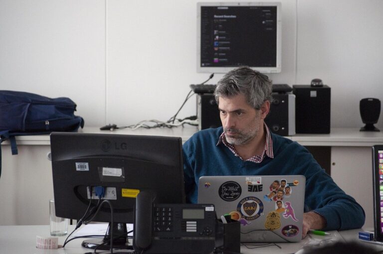 Middle-aged man with gray hair and beard wearing blue sweater working on laptop computer at modern office desk