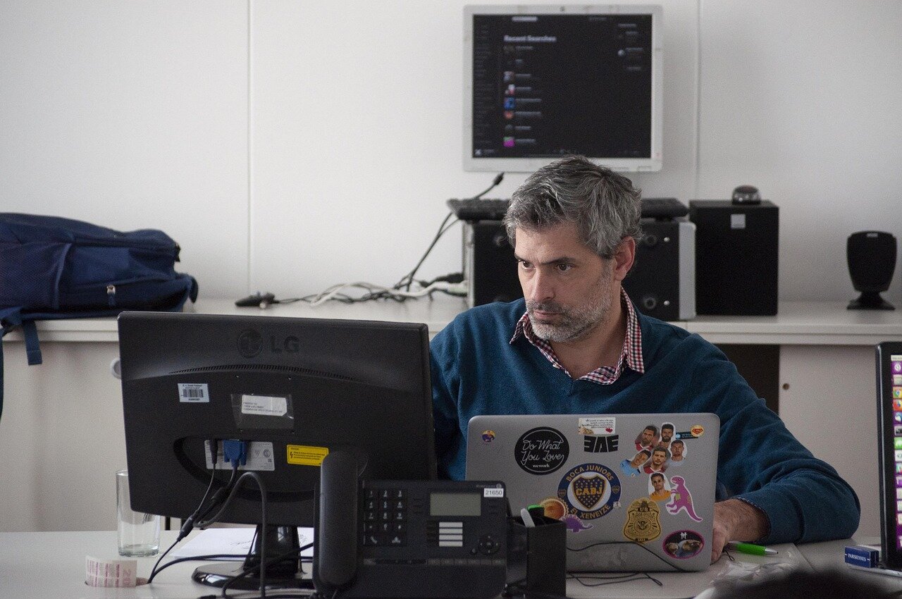 Middle-aged man with gray hair and beard wearing blue sweater working on laptop computer at modern office desk
